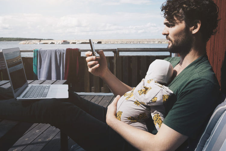 Father with laptop holding baby boy while using mobile phone at holiday villa