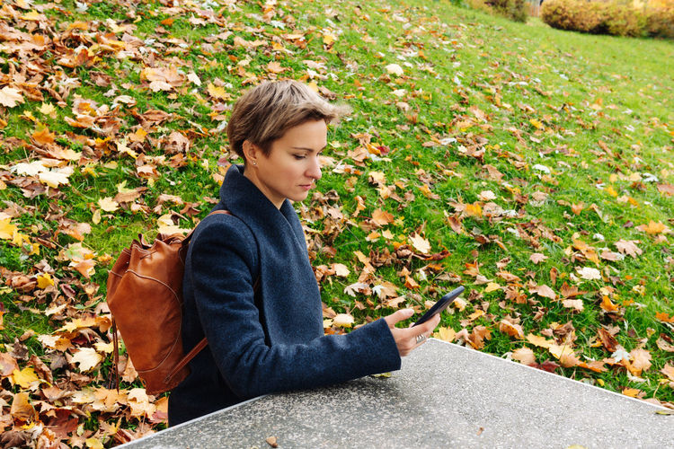 A woman looks at the screen of her phone sitting at a table in an autumn park.
