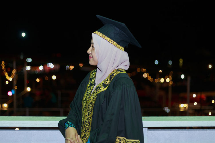 Smiling young woman wearing mortar board while standing against sky at night