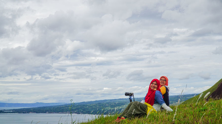 Female friends sitting on grassy hill by lake toba against sky