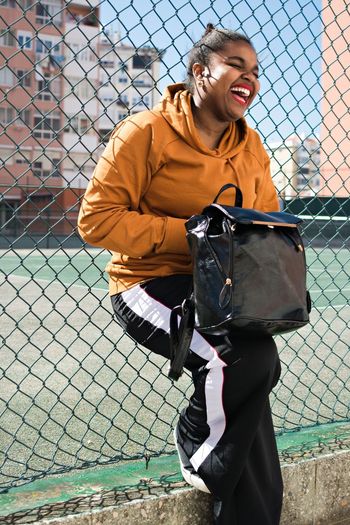 Smiling young woman with bag leaning on chainlink fence
