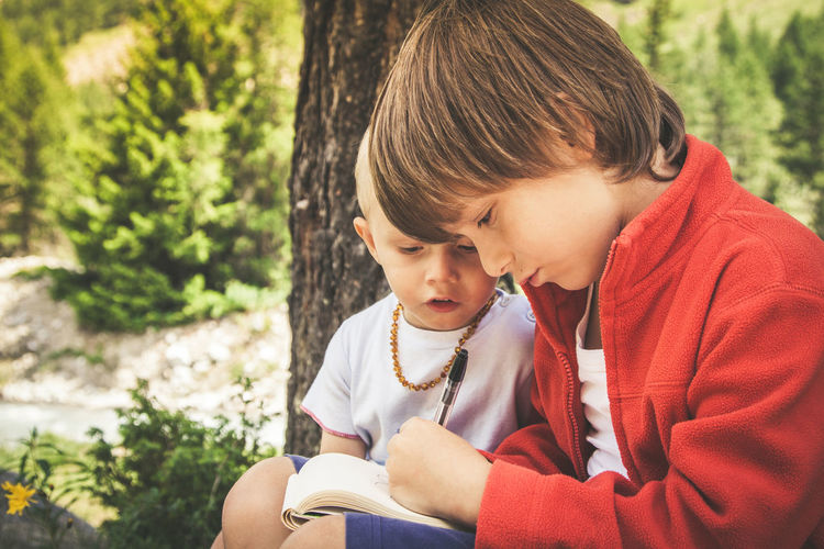 Cute boy looking at brother writing in book while sitting outdoors