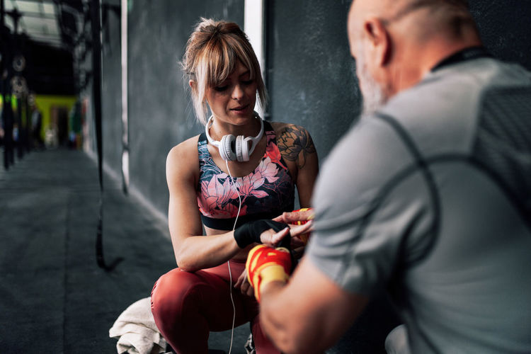Adult bearded male instructor helping woman athlete wrapping bandage on wrist while sitting near black wall and preparing for exercising in boxing gym