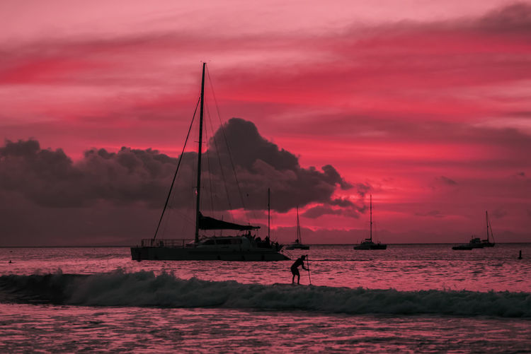 Silhouette man paddleboarding by boats on sea against dramatic sky