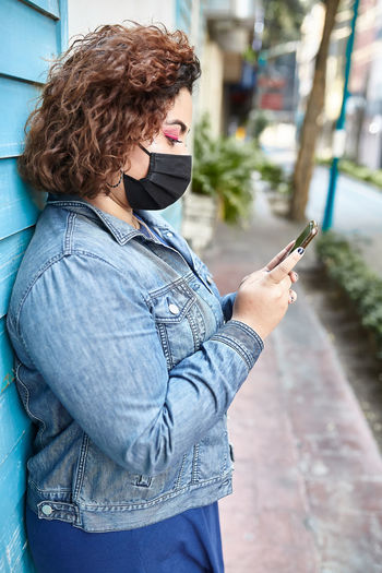 Side view of plus size female wearing protective mask standing in narrow street near residential building and browsing smartphone during covid 19 epidemic in city