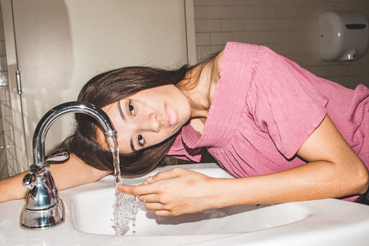 Portrait of teenage girl lying on sink with running tap water in bathroom at home