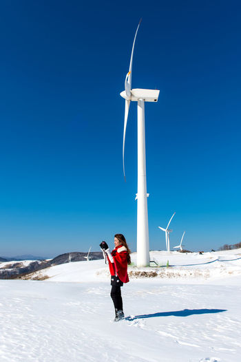 Woman holding camera while standing on snow covered road against wind turbines and clear blue sky