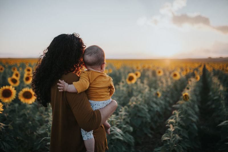 Mother carrying toddler daughter in sunflower farm against sky