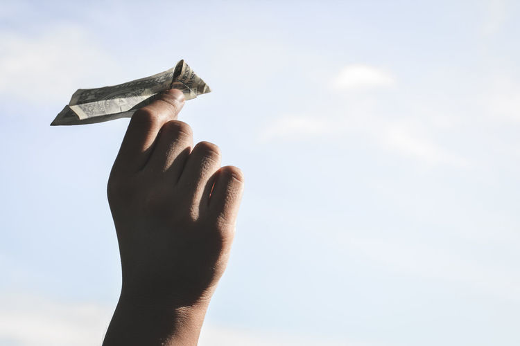 Cropped image of hand holding paper currency airplane against sky