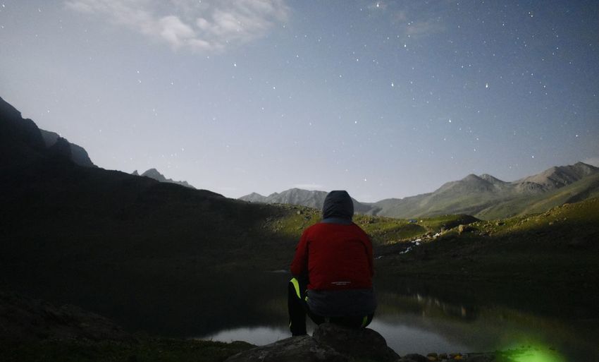 Rear view of man looking at mountain while sitting by lake against sky