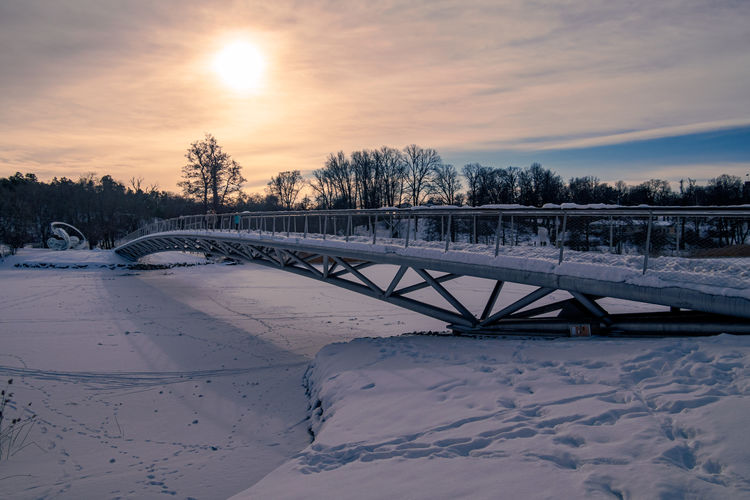 Bridge over snow covered field against sky during sunset