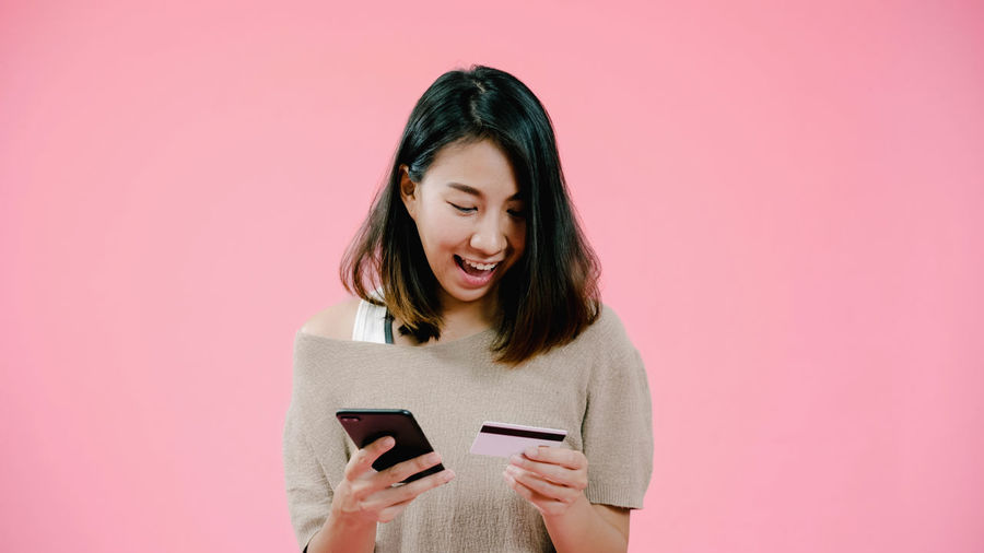Smiling woman doing online shopping through mobile phone against pink background
