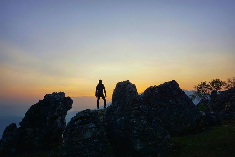 Rear view of man standing on cliff against sky during sunset