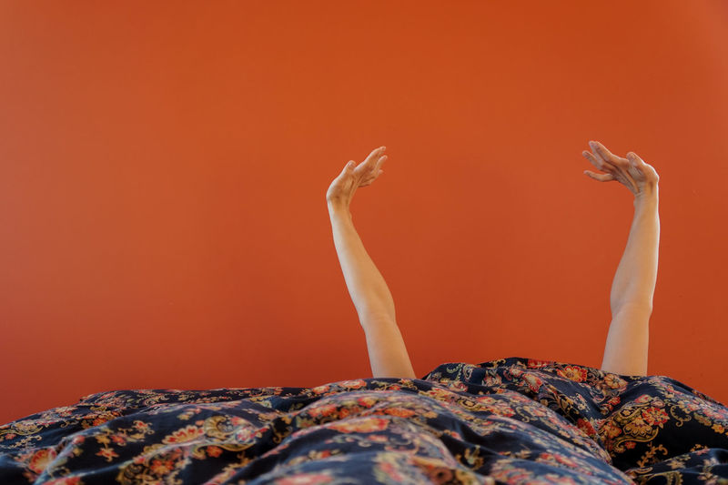 Cropped hands of person by blanket on bed against orange wall