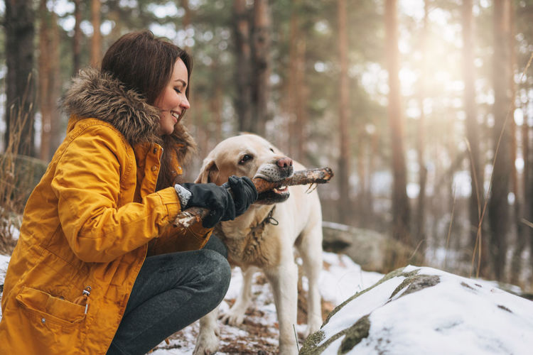 Young woman in yellow jacket trains big kind white dog labrador walking in winter forest