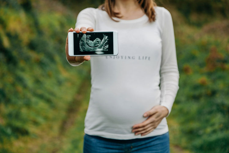 Midsection of woman holding mobile phone with ultrasound while standing on grass