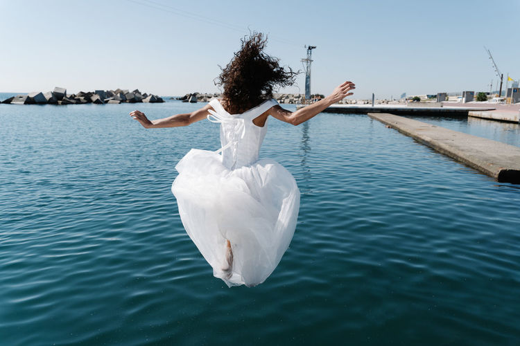 Back view of unrecognizable young female in white wedding dress falling in rippling lake near pier