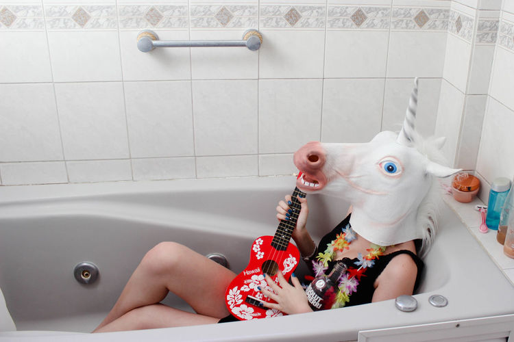 Woman wearing horse mask sitting with guitar in bathtub at home