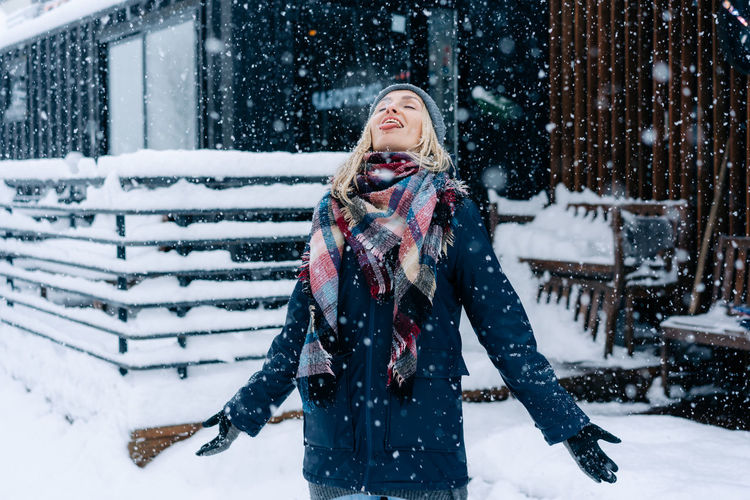 A blonde woman standing under a snowfall catches snowflakes with her tongue like a child having fun.