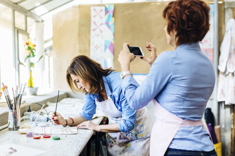 Woman taking picture of partner drawing on fabric in workshop