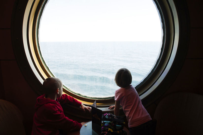Siblings looking at sea while standing by porthole in cruise ship