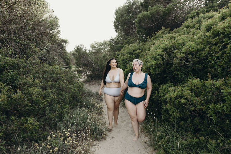 Positive curvy female friends in swimwear walking on sandy path on beach and enjoying summer