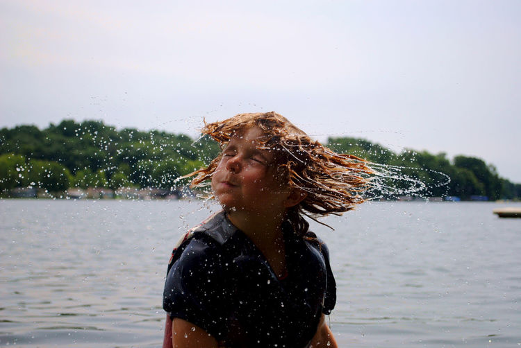 Boy tossing wet hair while standing at lake against sky