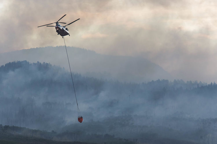 Military helicopter flying with fire retardant while smoke emitting from wildfire during sunset