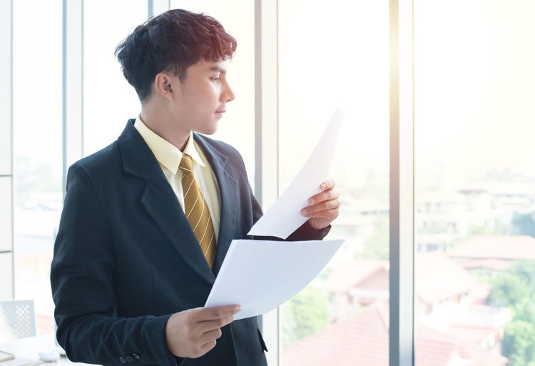 Young businessman holding document while standing by window in office