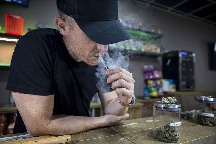 Anonymous adult male in cap smoking cannabis joint in workspace on blurred background