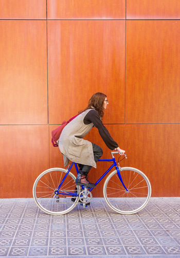 Pretty young woman in eyeglasses riding bicycle against red wall.