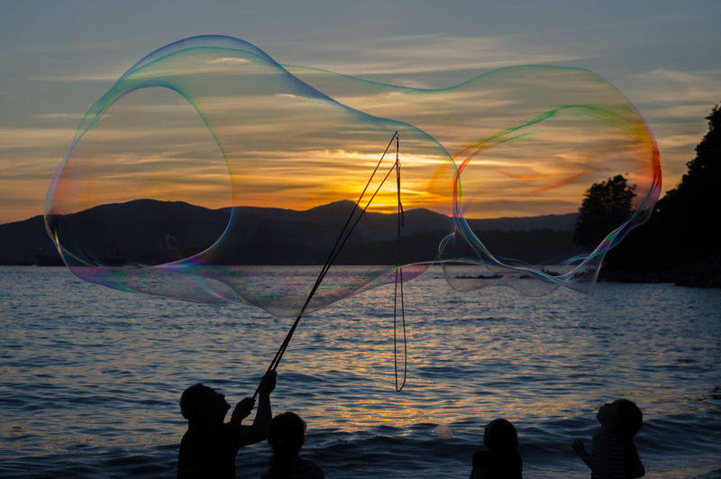 Silhouette man making huge bubbles at beach against sky during sunset