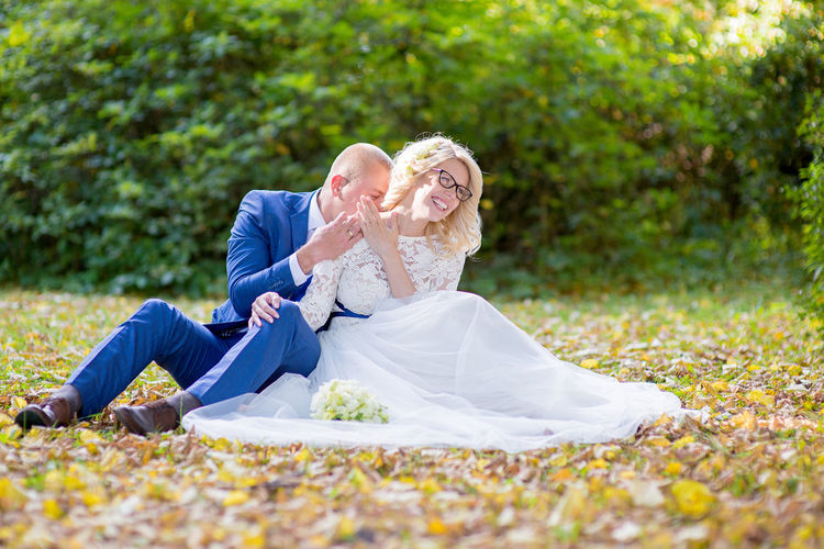 Smiling bride with groom sitting on field against plants at park