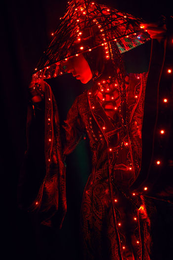 Enigmatic female in creative traditional outfit and vietnamese headwear with red illumination standing in dark studio on black background during performance