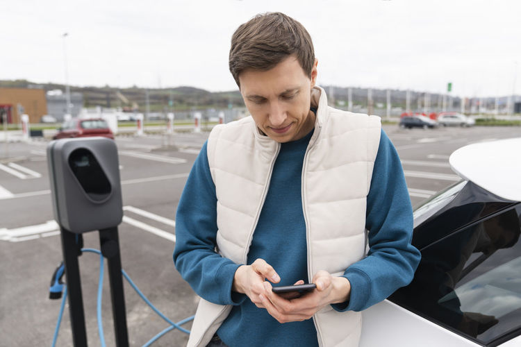 Man using smart phone leaning on car at charging station