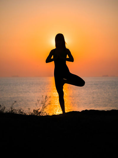 Silhouette woman doing tree pose while exercising on beach against sky during sunset