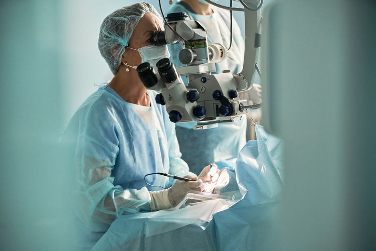 Focused adult female doctor in sterile mask and ornamental medical cap looking through surgical microscope against crop coworker in hospital