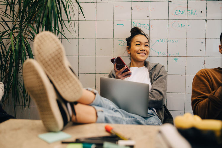 Smiling young businesswoman sitting with feet up on desk using wireless technologies by colleague against wall at office