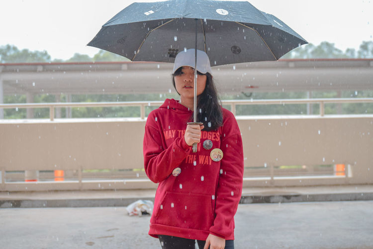 Young woman holding umbrella while standing on road during rainfall in city
