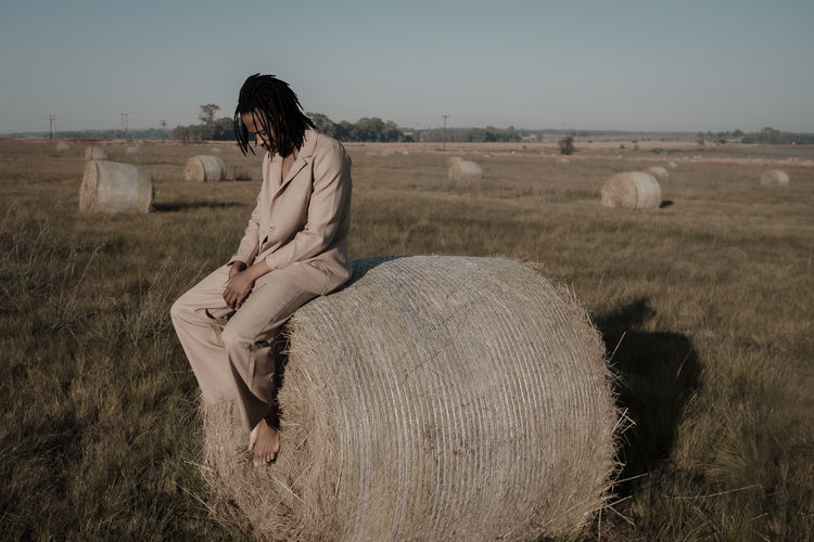 Full length of young man sitting on hay bale at agricultural landscape
