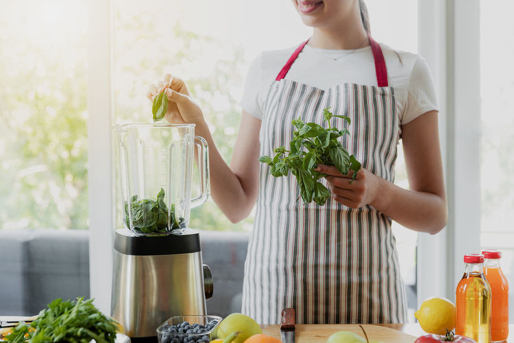 Young smiling woman cooking green spring smoothie at home kitchen using electronic blender device