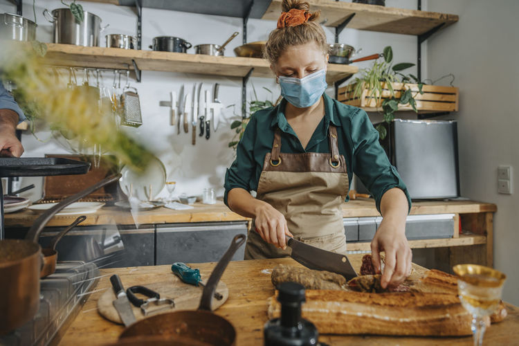 Young chef wearing protective face mask cutting salami while standing in kitchen