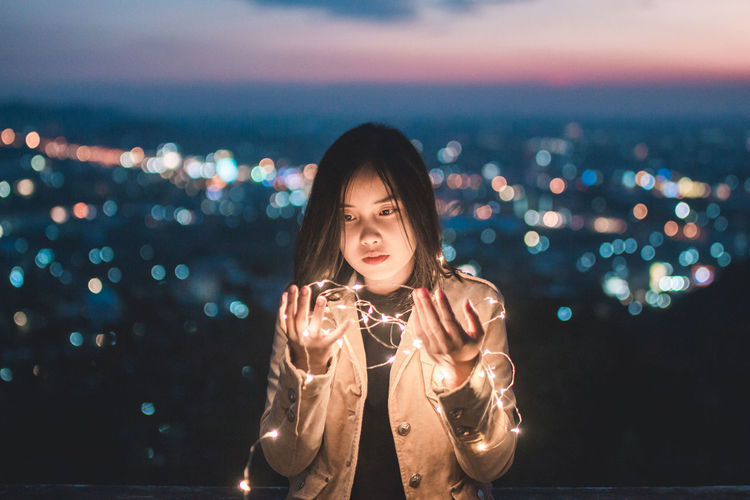 Young woman with illuminated string lights standing against illuminated cityscape at night
