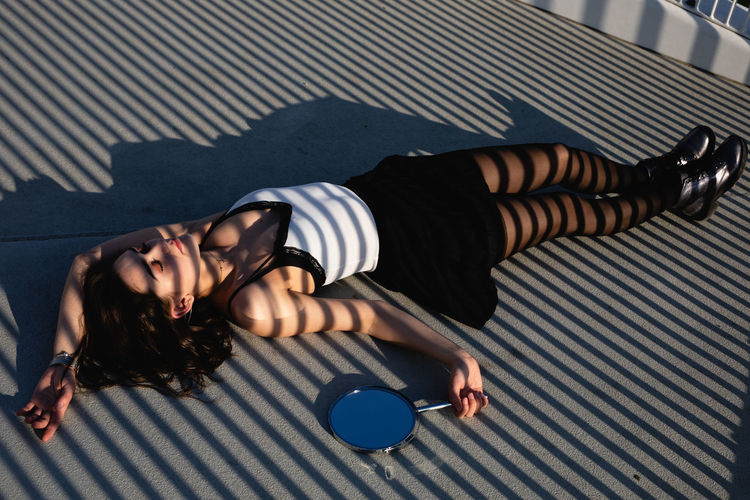 Full length of young woman holding hand mirror while lying down on elevated walkway
