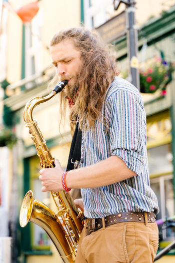Vertical photo of a young man with long hair playing saxophone in the street