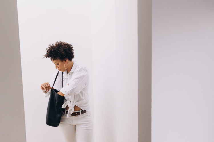 Portrait of young man standing against a white wall, looking for in her bag