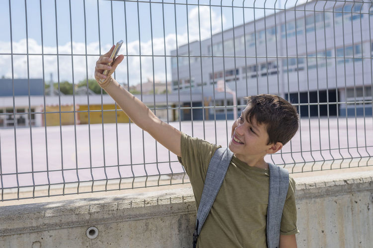 Smiling boy taking selfie through mobile phone while standing against wall