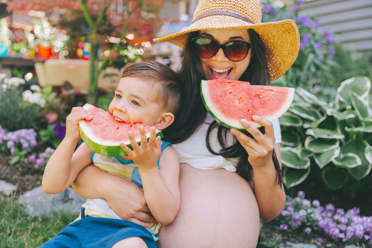 Pregnant mother and son eating watermelons while sitting against plants