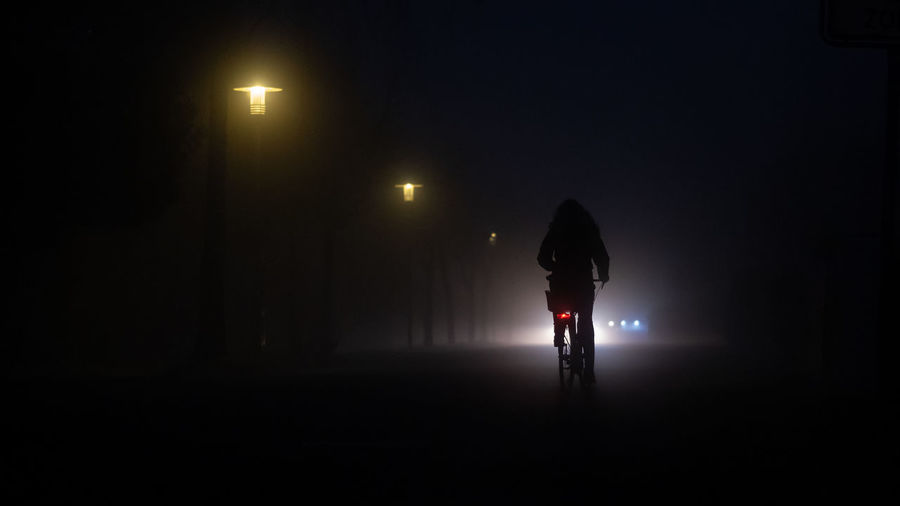 Rear view of dark silhouette person on bicycle on street at night