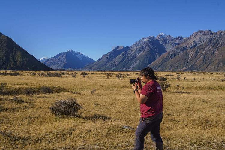 Side view of man photographing with digital camera while standing on field against mountains